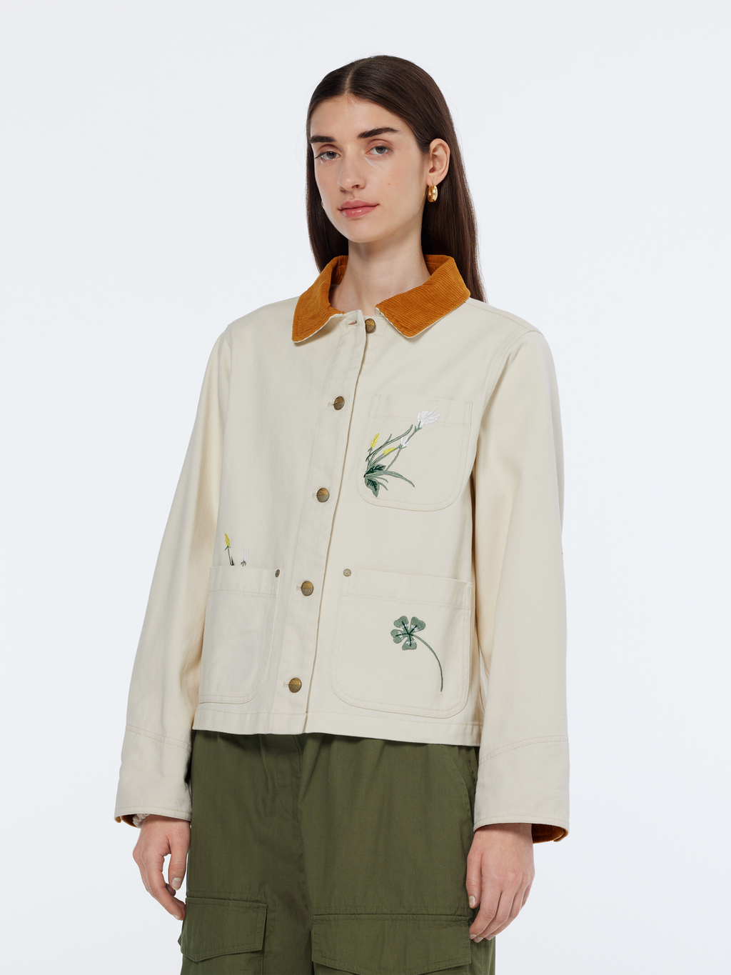 A woman wearing a beige embroidered barn jacket with a colorful collar and button-up style, paired with green pants.
