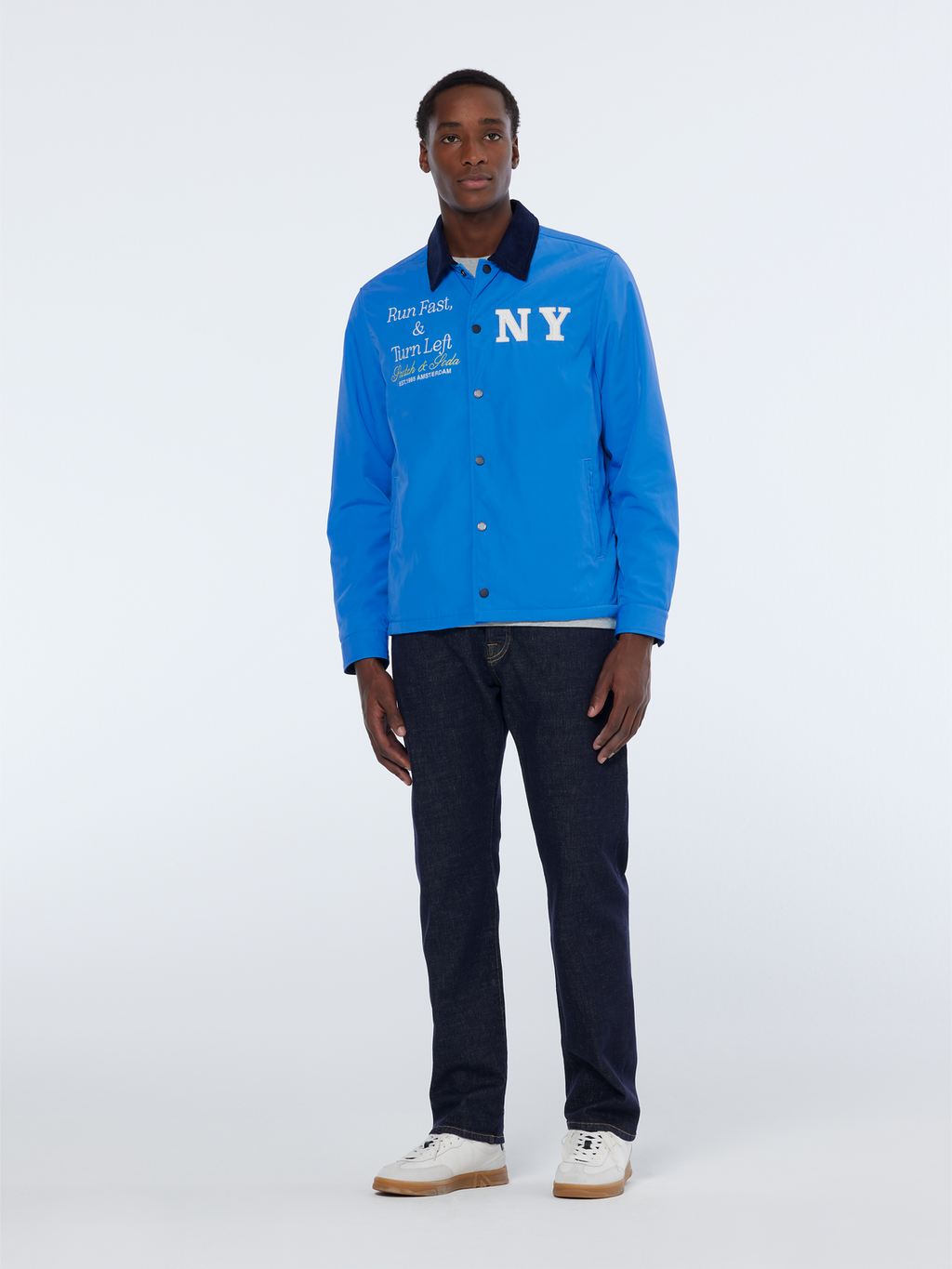 A man wearing a blue satin club jacket with 'NY' text on the front, paired with dark trousers and white shoes.