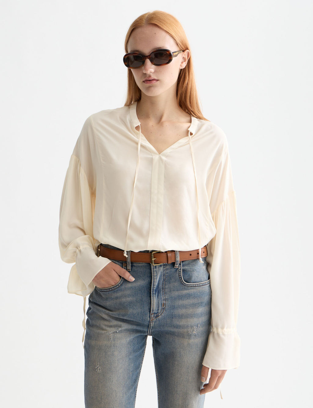 Woman wearing a cream blouse and blue jeans with sunglasses on a white background