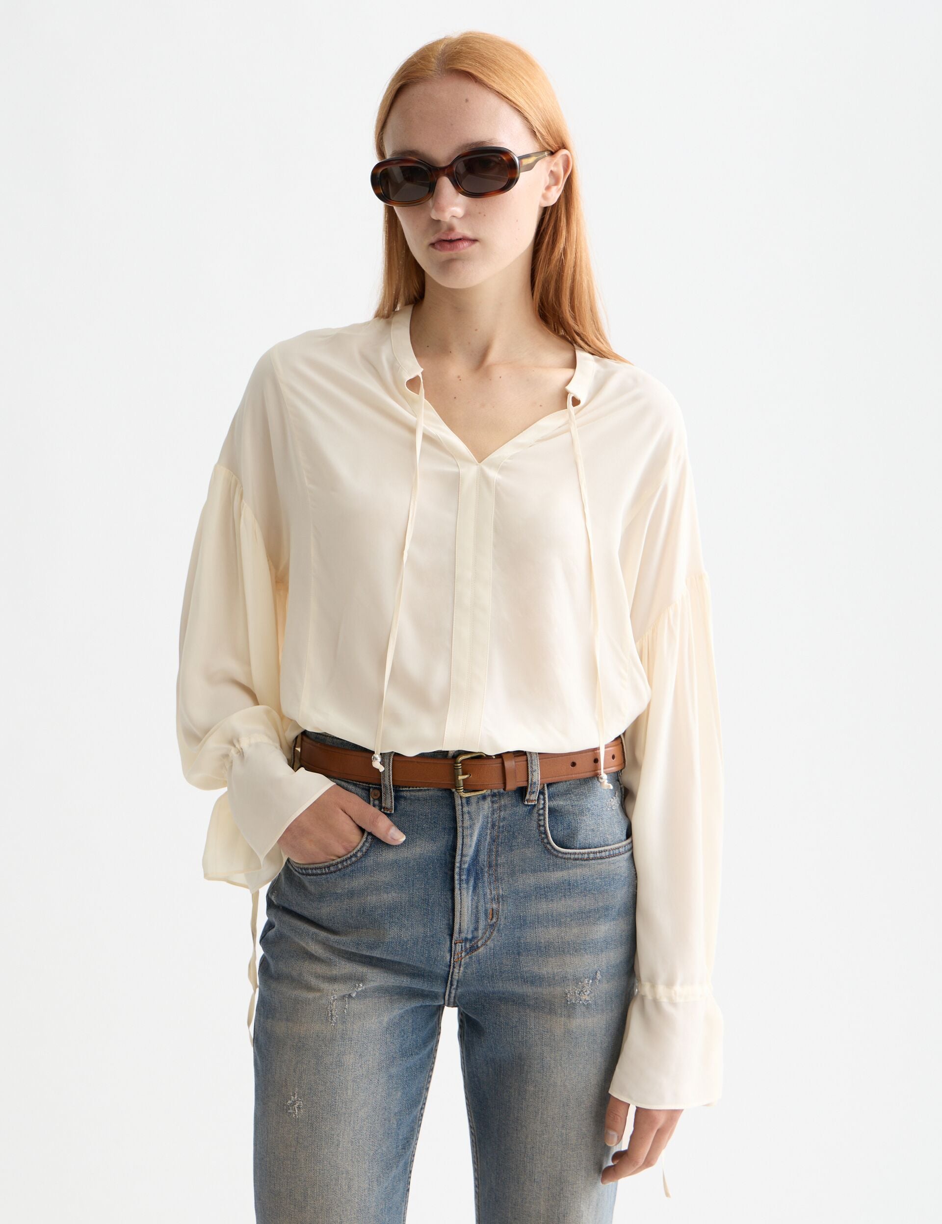 Woman wearing a cream blouse and blue jeans with sunglasses on a white background