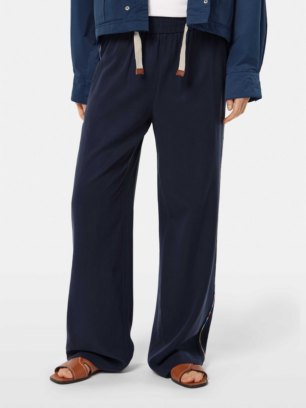 A model wearing navy mid-rise wide-leg trousers with side zip details, paired with a blue top and brown sandals.